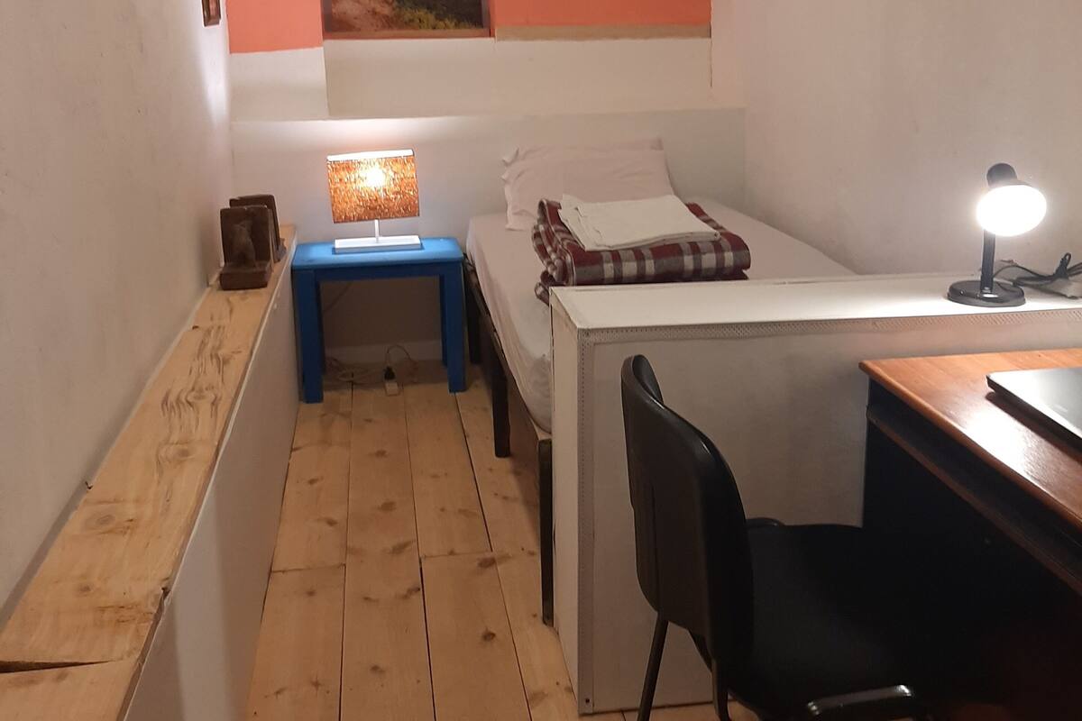 A compact room with a bed, a desk, a chair, a small table, and a wall-mounted lamp.