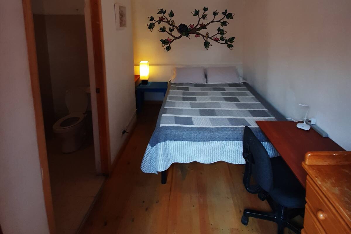 A small, well-lit bedroom with a single bed, a desk, and a dresser. There is a wall-mounted lamp and a ceiling light fixture.