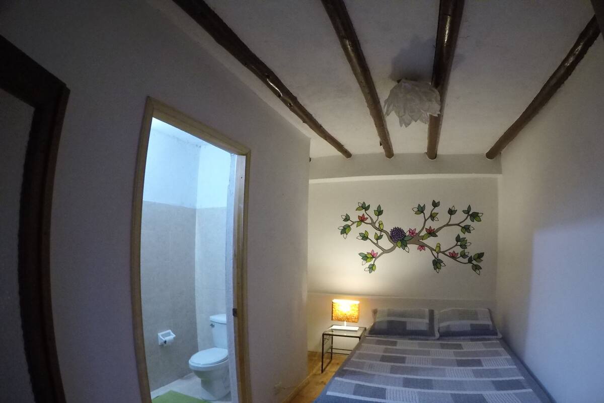A bedroom with a bed, a nightstand, a lamp, a wooden floor, a ceiling with exposed beams, and a bathroom visible through an open door.