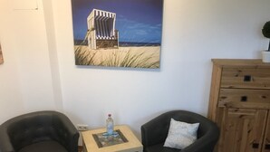 Una habitación con dos sillones negros, una pequeña mesa de madera con una botella, y un cuadro de una silla de playa en la pared.