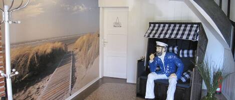 A room with a mural of a beach scene, a coat rack, a door, a chair with a checkered awning, and a vase with plants.