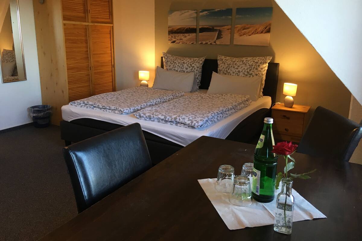 A bedroom with a bed, two bedside lamps, a nightstand, a table with a bottle and glasses, and a painting on the wall.