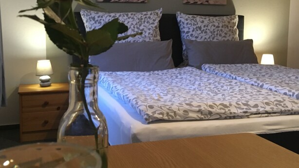 Habitación de hotel con cama, mesitas de noche, un jarrón con una rosa roja y dos cuadros colgados.