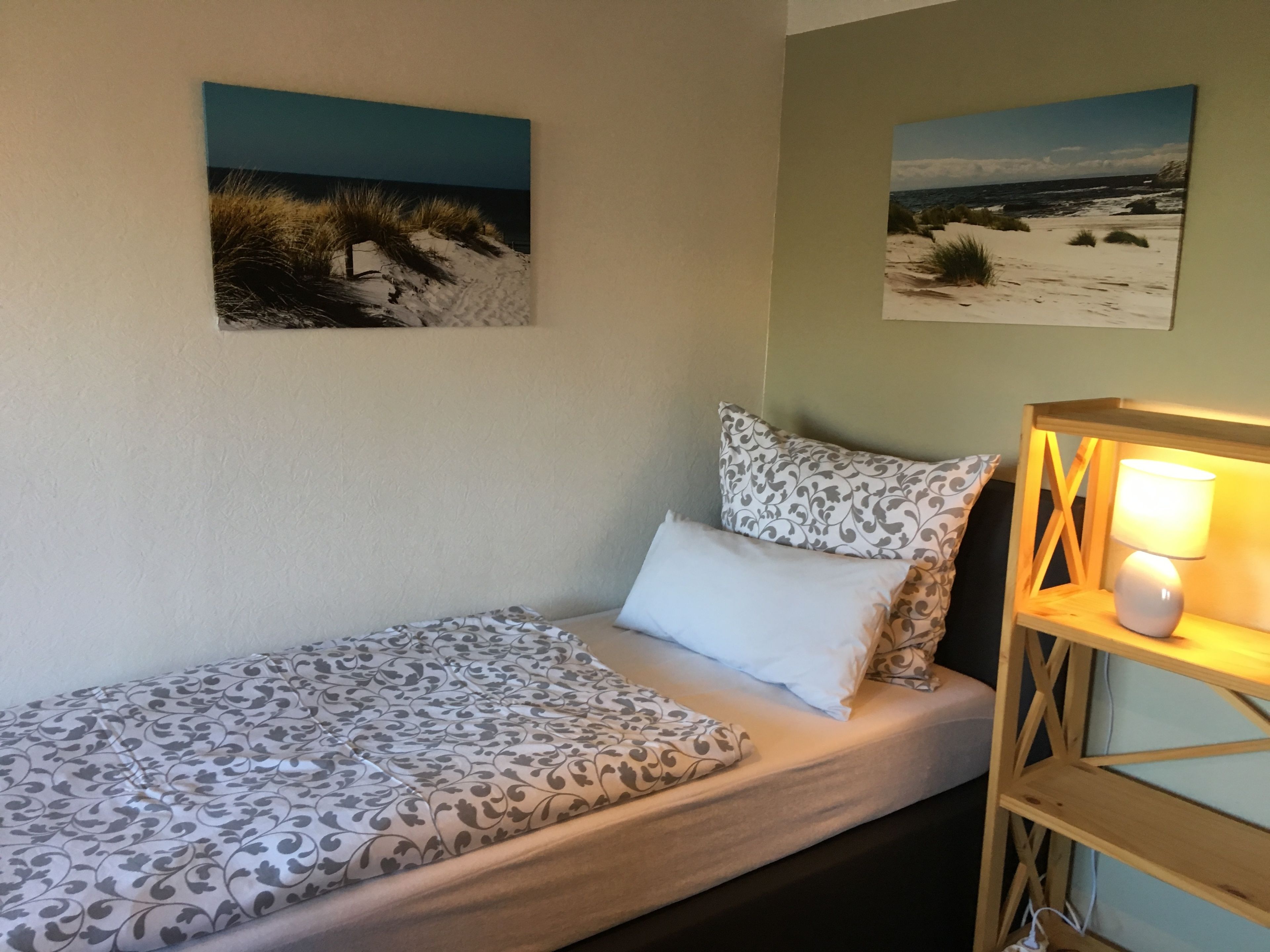 A single bed with patterned bedding, a bedside lamp, and two wall-mounted paintings of beach scenes.