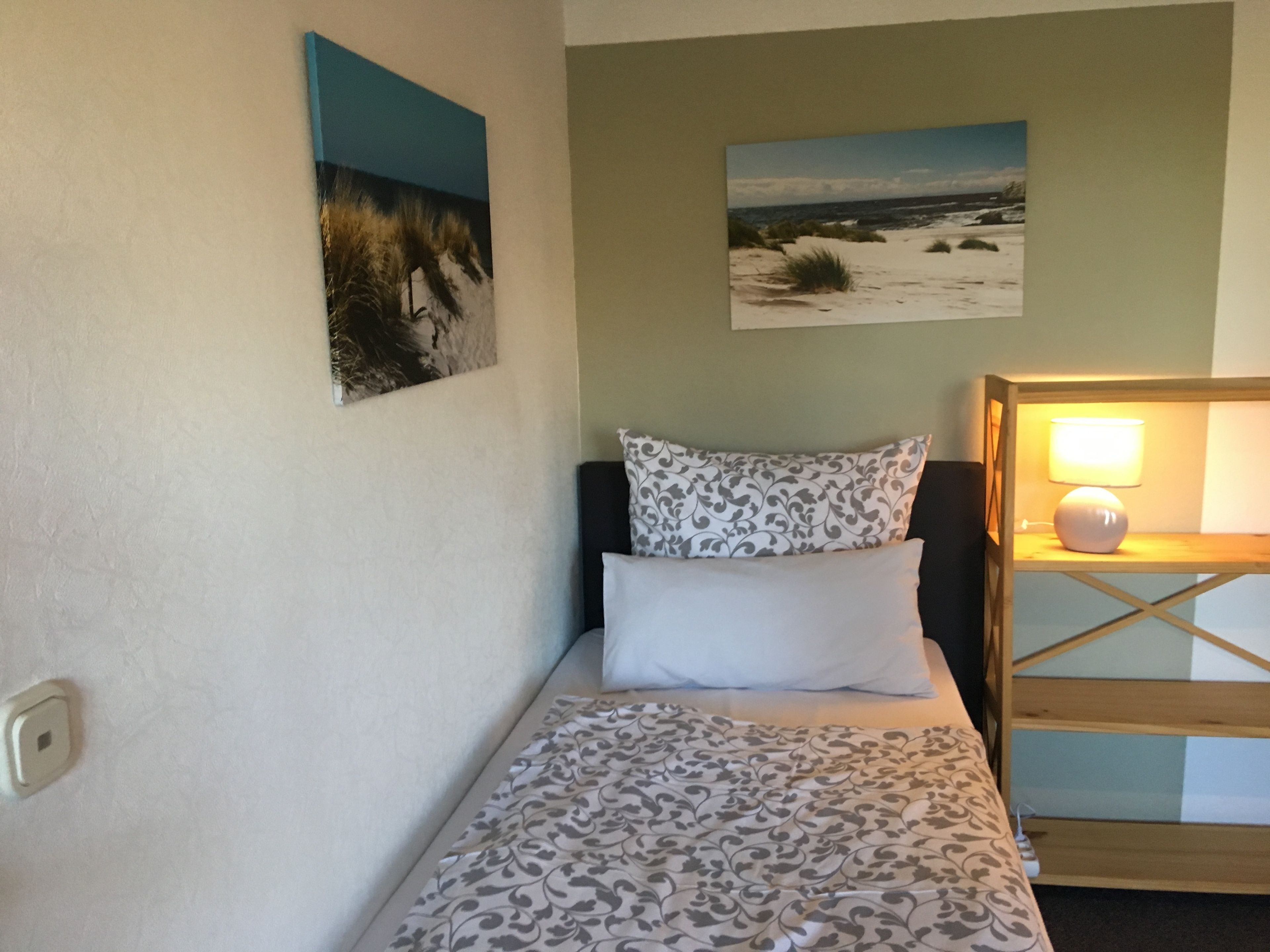 A single bed with patterned bedding, a nightstand with a lamp, and two wall-mounted pictures depicting landscapes.