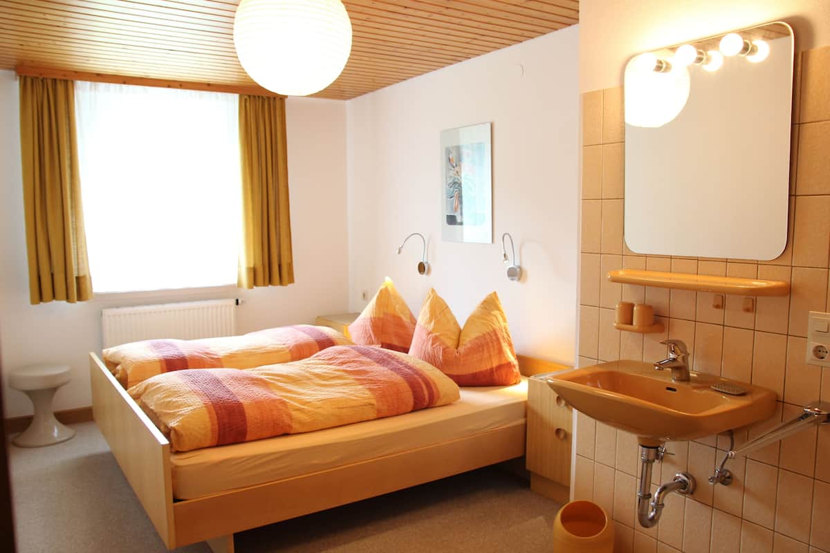 Quarto com cama de madeira, mesa de cabeceira, espelho e banheiro com pia e vaso sanitário.
