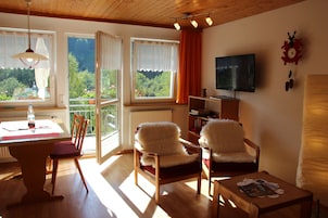 Una sala de estar acogedora con techo de madera, una mesa de comedor con sillas, un sofá, un televisor y vistas al verdor exterior.