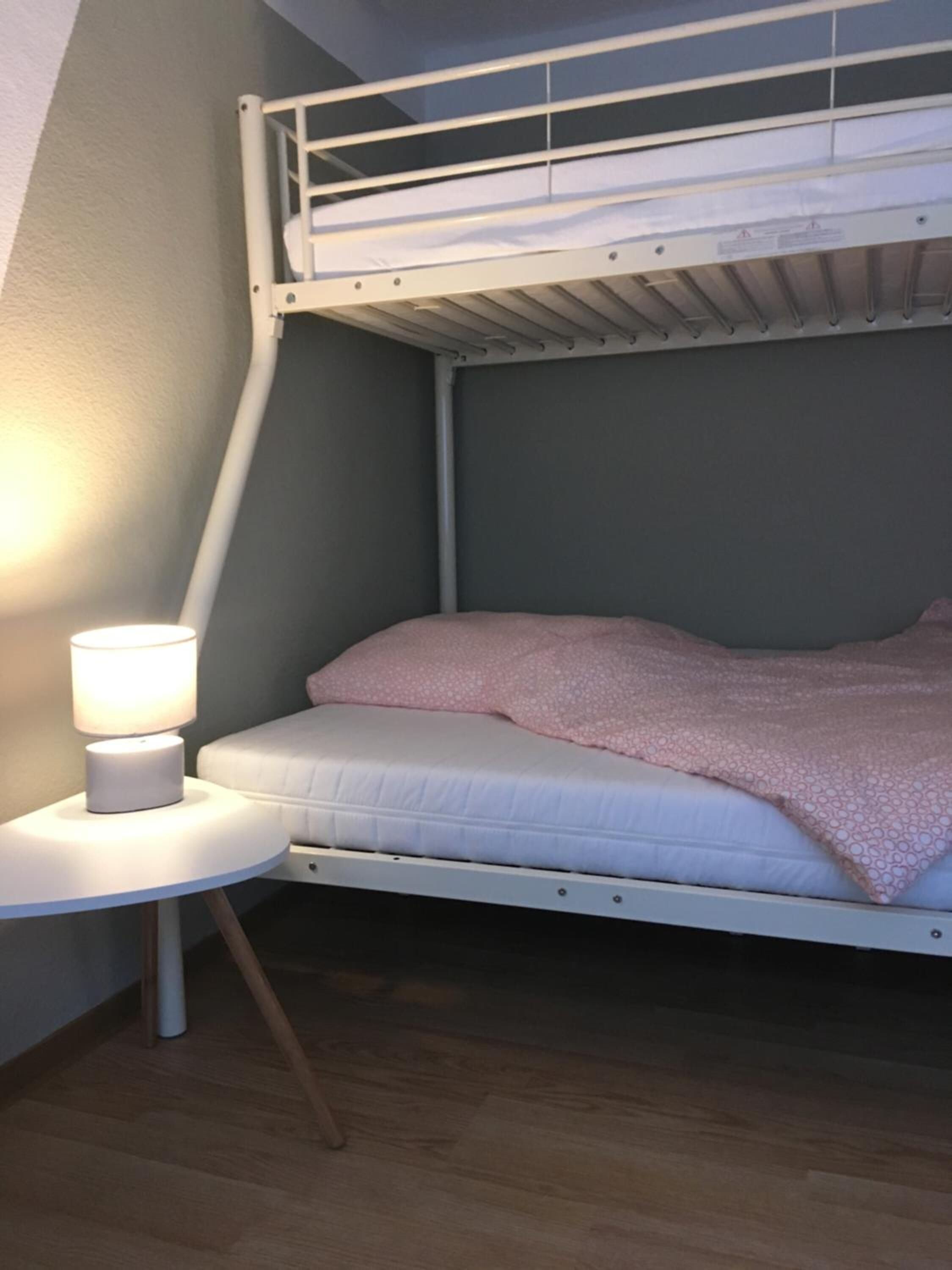 Ein Stockbett mit weißem Gestell, ein runder Nachttisch mit Lampe und eine pinkfarbene, gemusterte Decke.