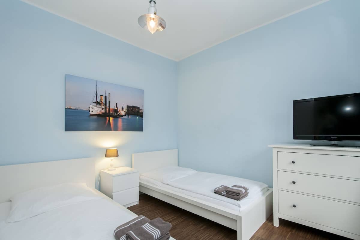 Habitación de hotel con cama, televisor, mesita de noche, lámpara y un cuadro de una escena portuaria.