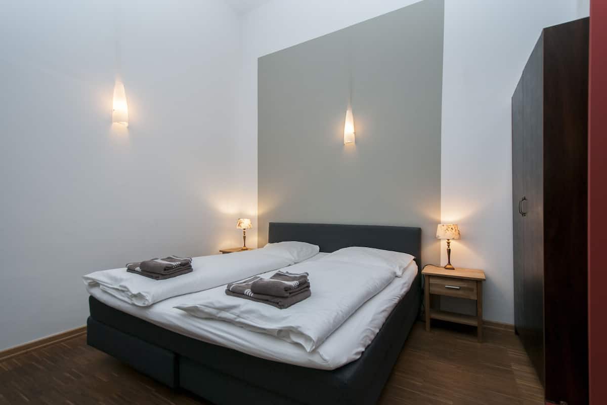 Una cama individual bien hecha con ropa blanca y dos toallas dobladas, mesitas de noche con lámparas y un armario de color oscuro.
