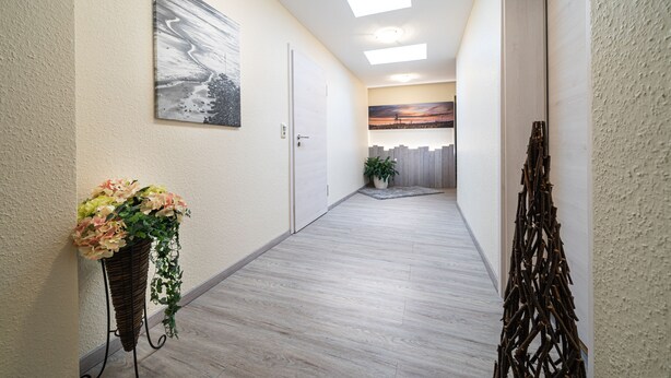 A hallway with a light wooden floor, a wall-mounted painting, a potted plant, and a decorative wall piece. - Deichhotel Grube (Wurster Nordseeküste)