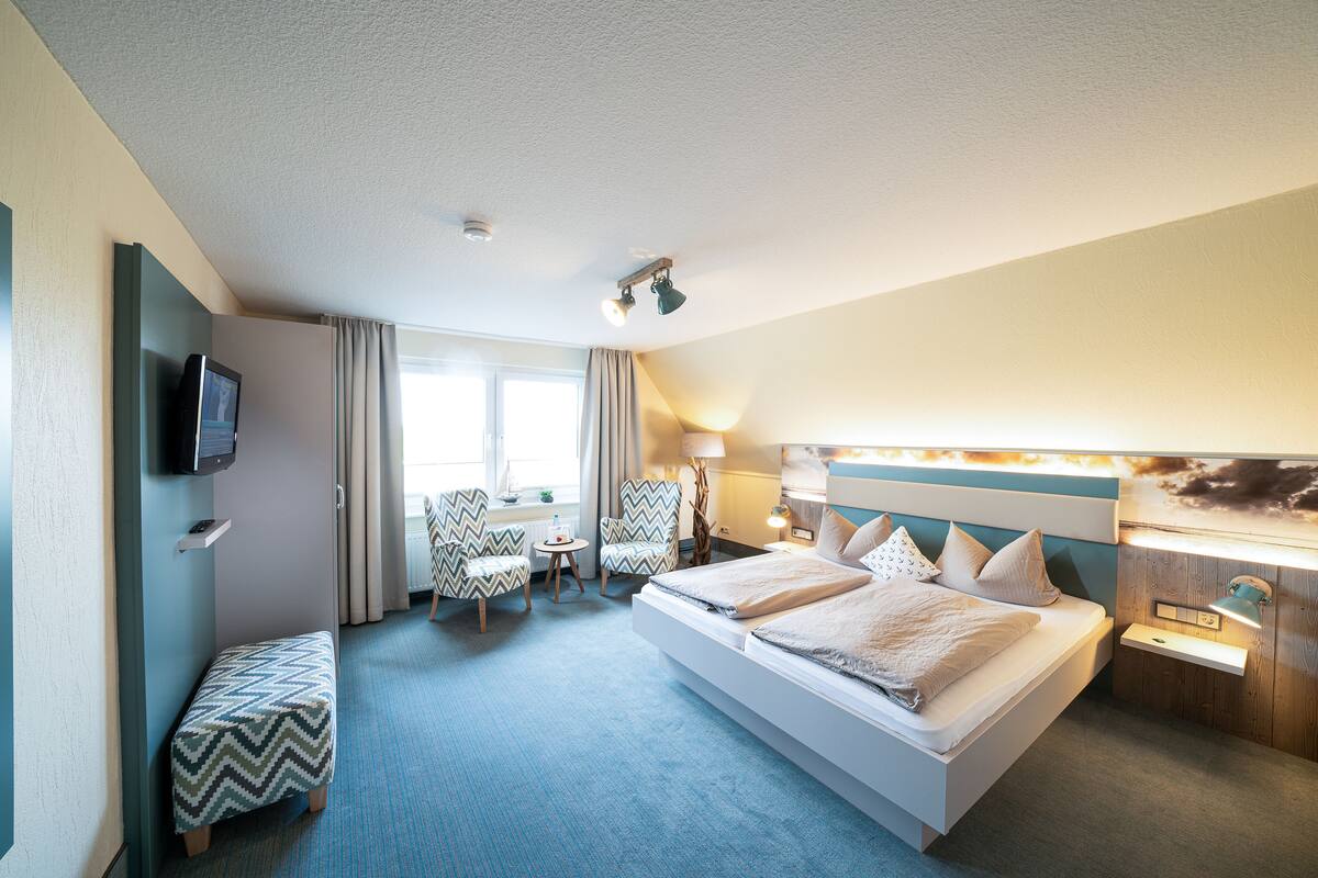 A hotel room with a large bed, two chairs, a small table, a TV, and a window with curtains.