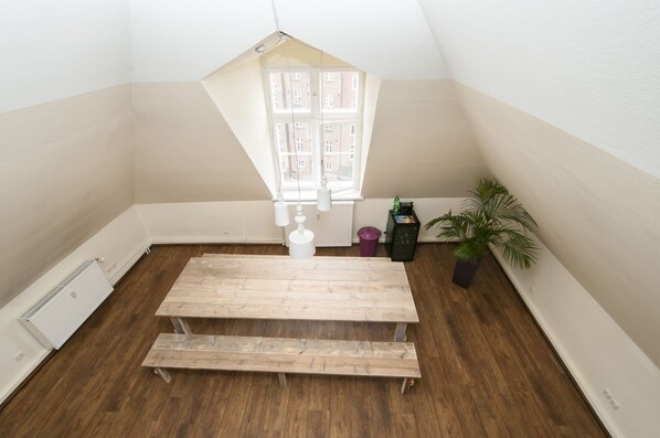 A small, well-lit room with a sloped ceiling, a wooden bench, a radiator, a potted plant, and a window with a view of buildings.