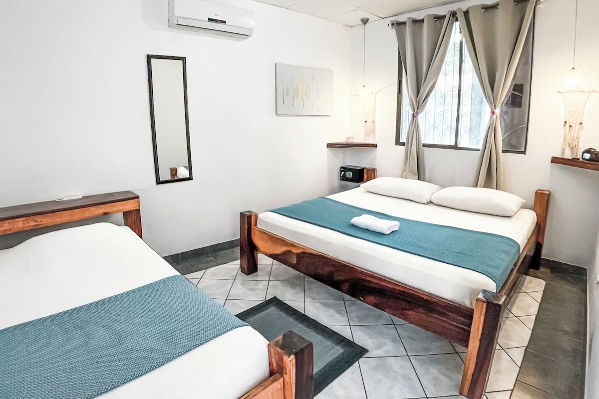 Habitación de hotel con dos camas, una mesita de noche, un aire acondicionado, una mesita con radio y una ventana con cortinas, además de una obra de arte colgada en la pared.