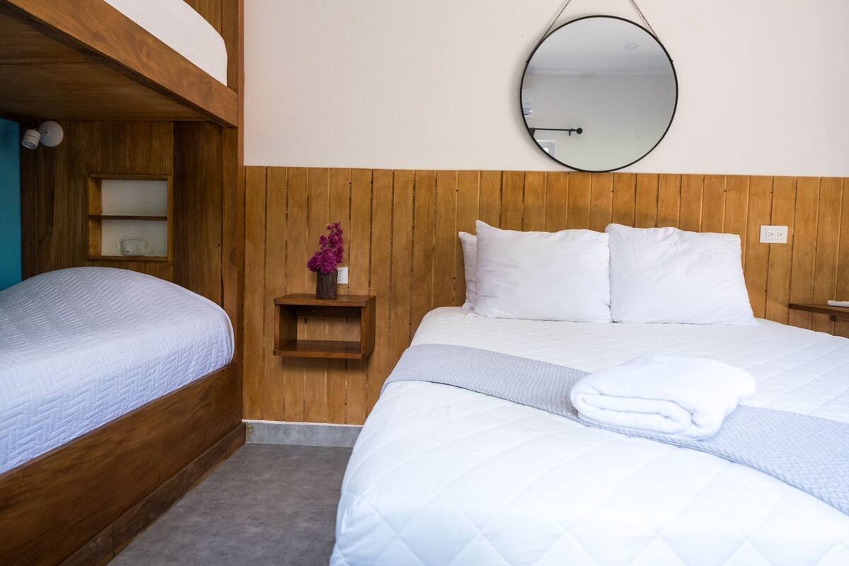 Una habitación de hotel con literas, una cama individual, un cabecero de madera, una mesita de noche con un florero y un espejo redondo en la pared.