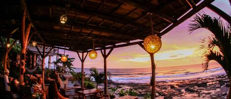 Un espacio al aire libre con mesas y bancos de madera, con vista a una playa al atardecer.