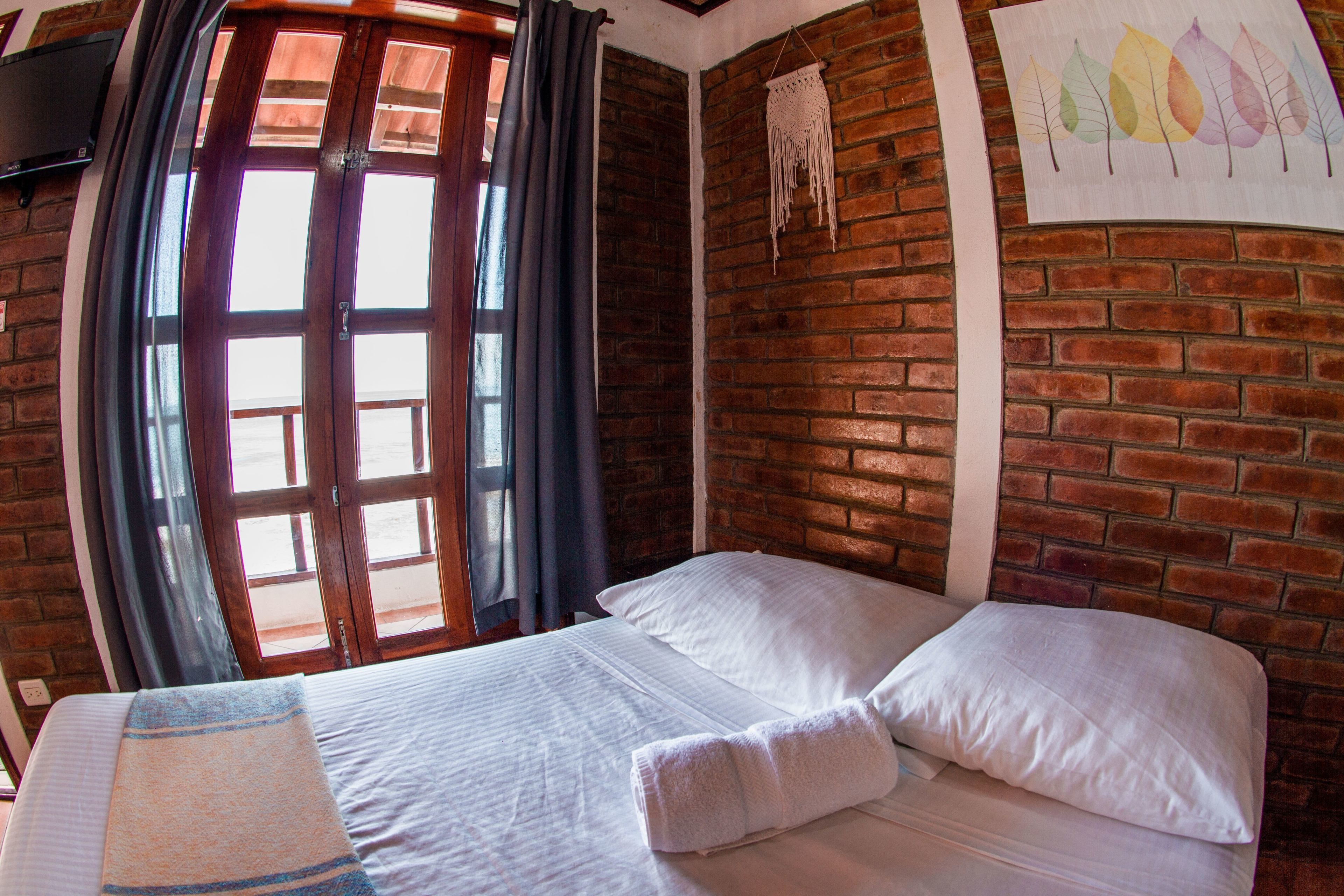 Een kamer met een bakstenen muur, een bed met witte lakens, een raam met gordijnen en een decoratie aan de muur.