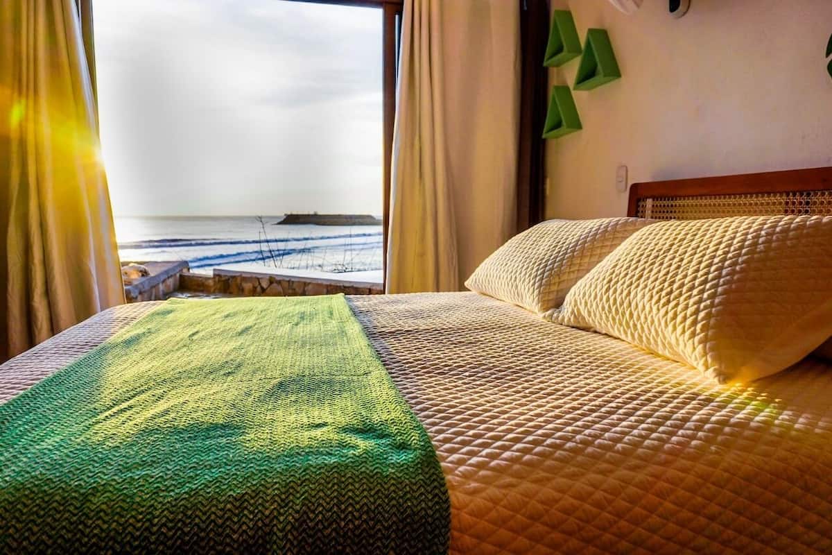 Una cama con un edredón verde y vistas a la playa a través de una ventana.