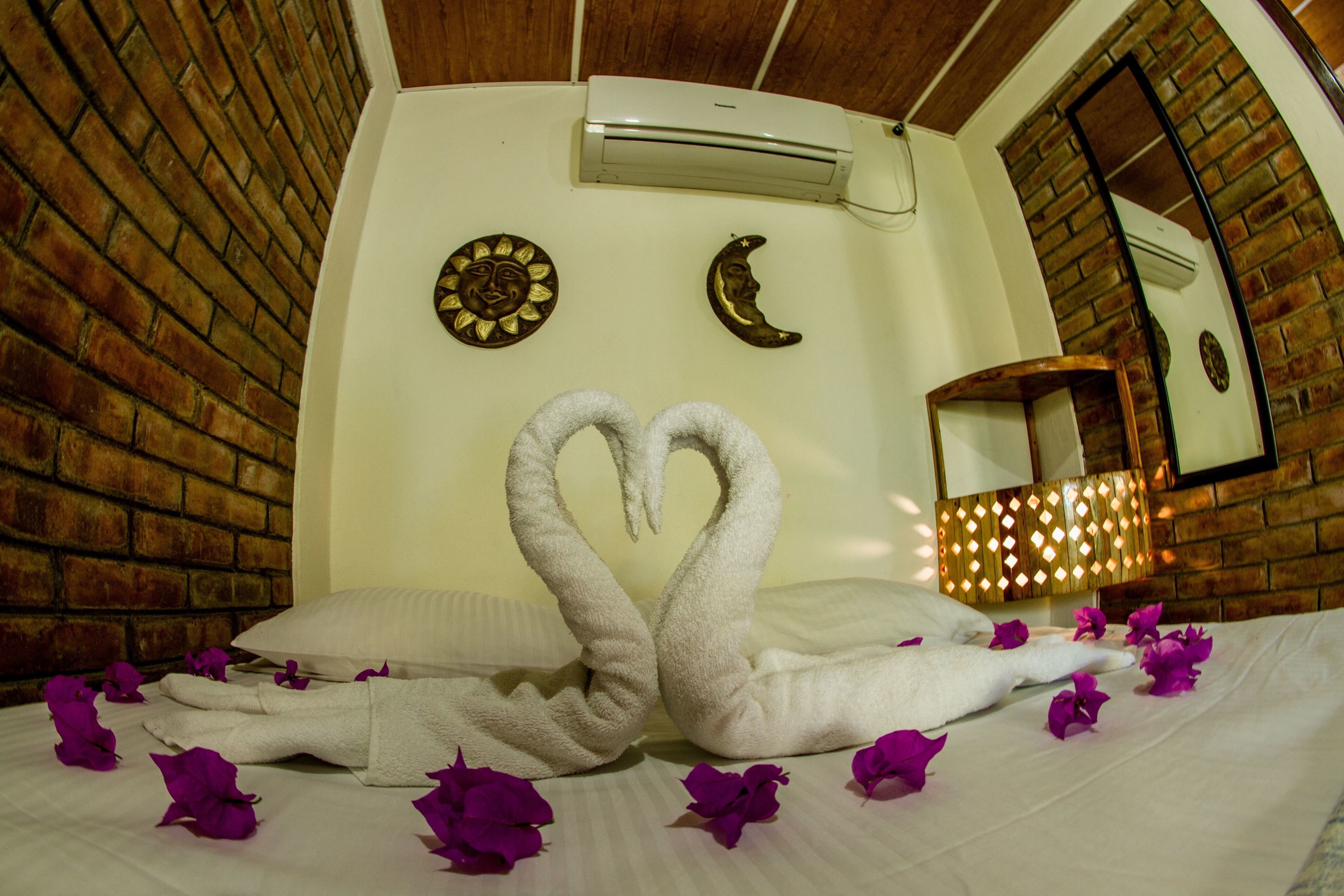 Una habitación de hotel con pared de ladrillo, una cama con ropa blanca, una toalla dispuesta en forma de corazón y una mesita de noche con una lámpara.