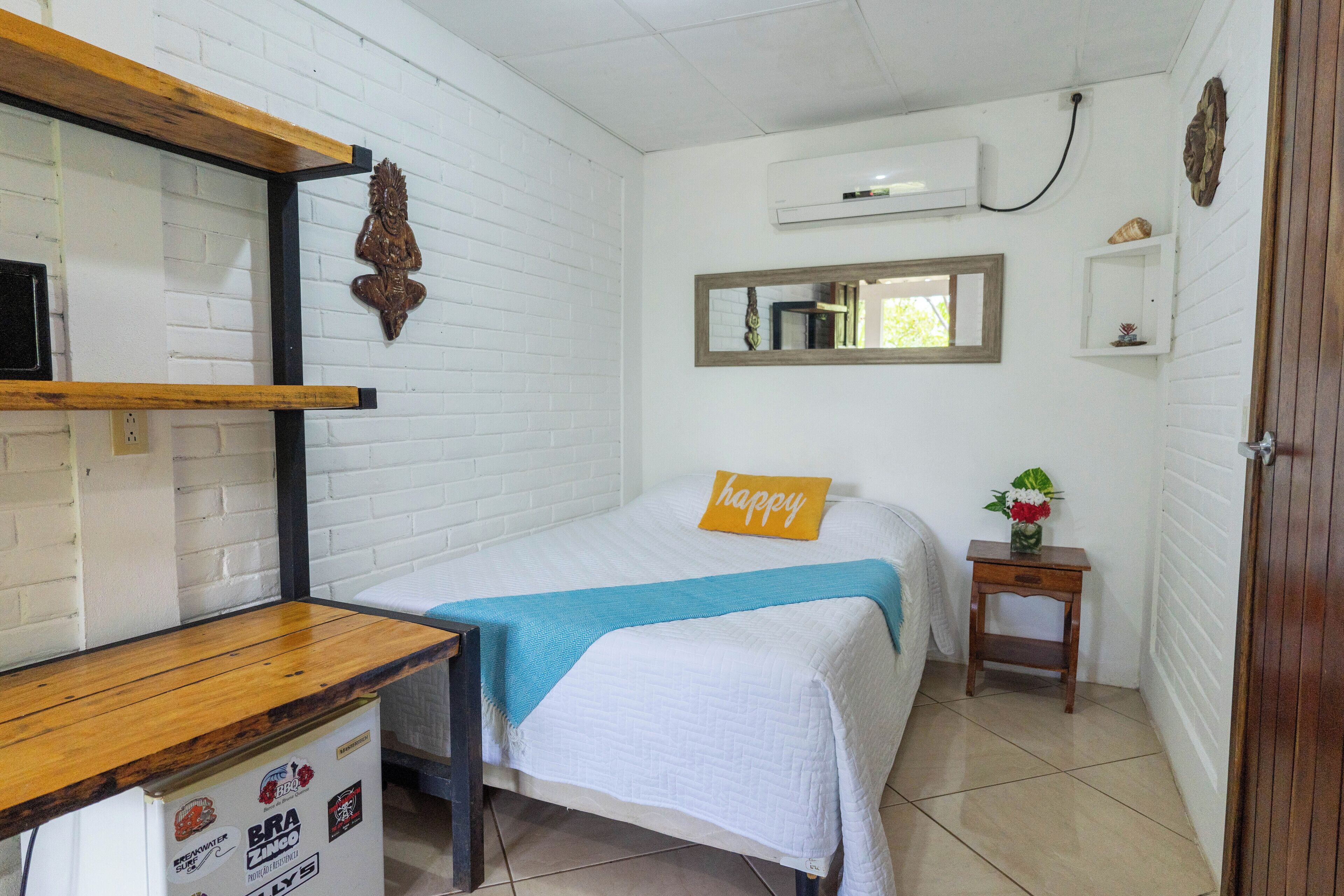 Um quarto pequeno e limpo com uma cama, uma escrivaninha de madeira, um espelho e uma pequena estante com objetos decorativos.