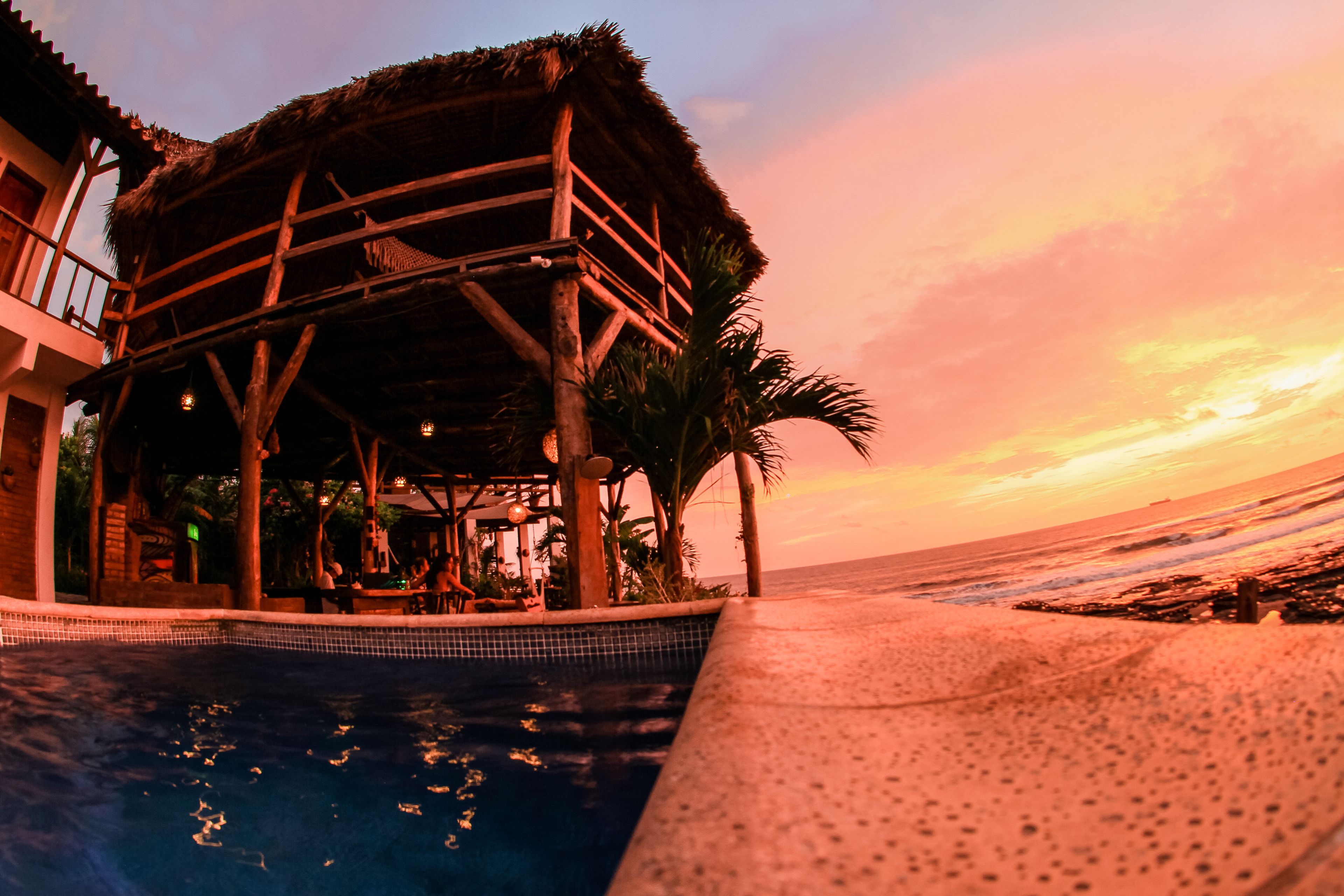 A poolside area with a wooden structure, palm trees, and a view of the ocean at sunset.
