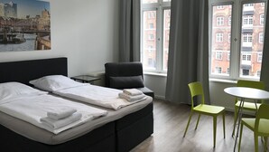 A hotel room with a bed, a chair, a table, and a window with a view of buildings. - Appartement 8er Deck (Hamburg)