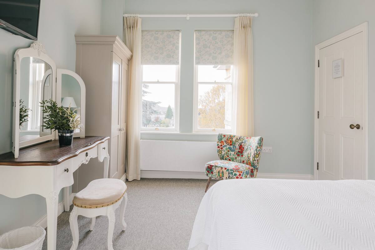 Une chambre à coucher avec un lit, une commode avec un miroir, une chaise et une fenêtre avec des rideaux.