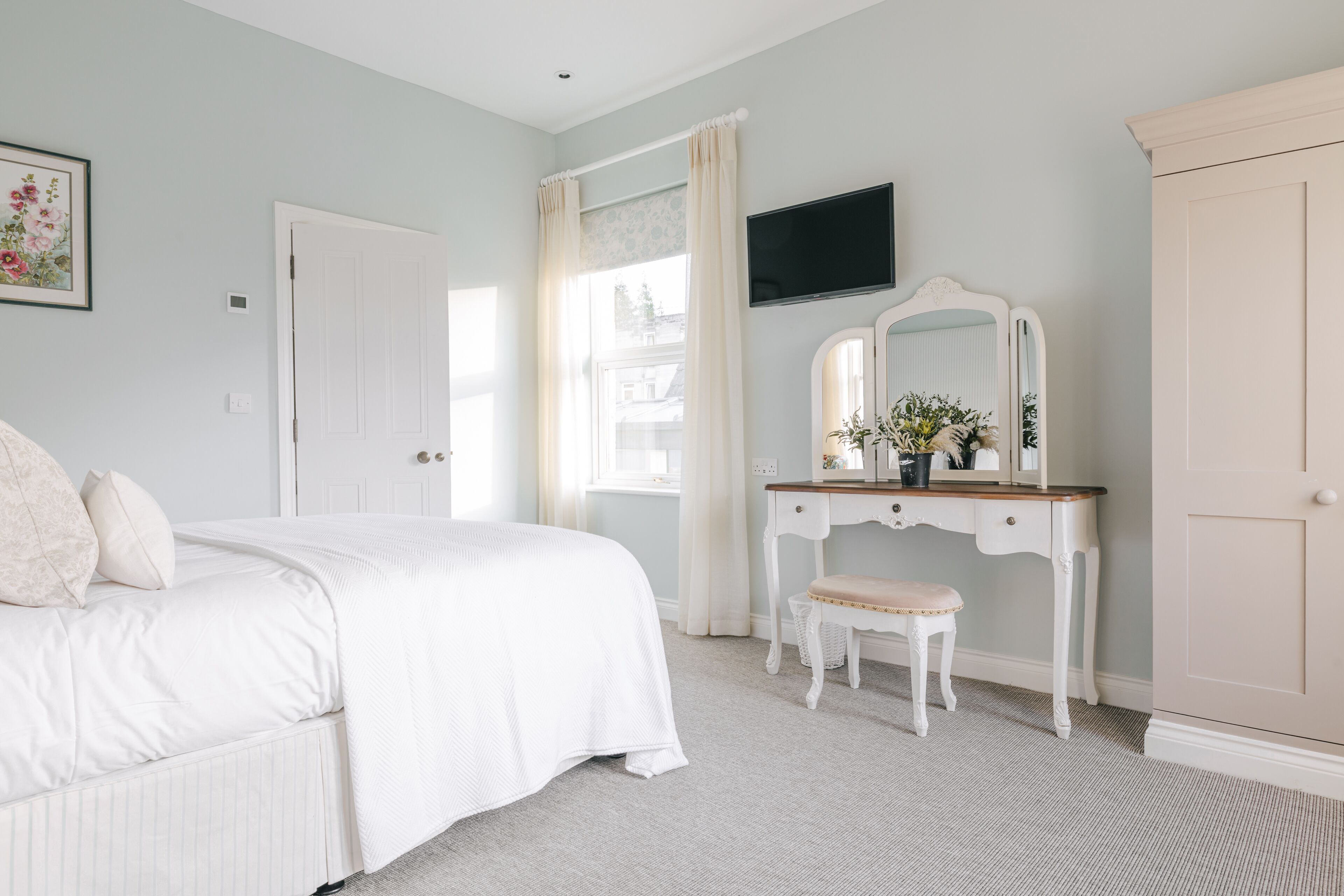 A bedroom with a bed, a vanity with a mirror, a dresser, and a television on the wall.