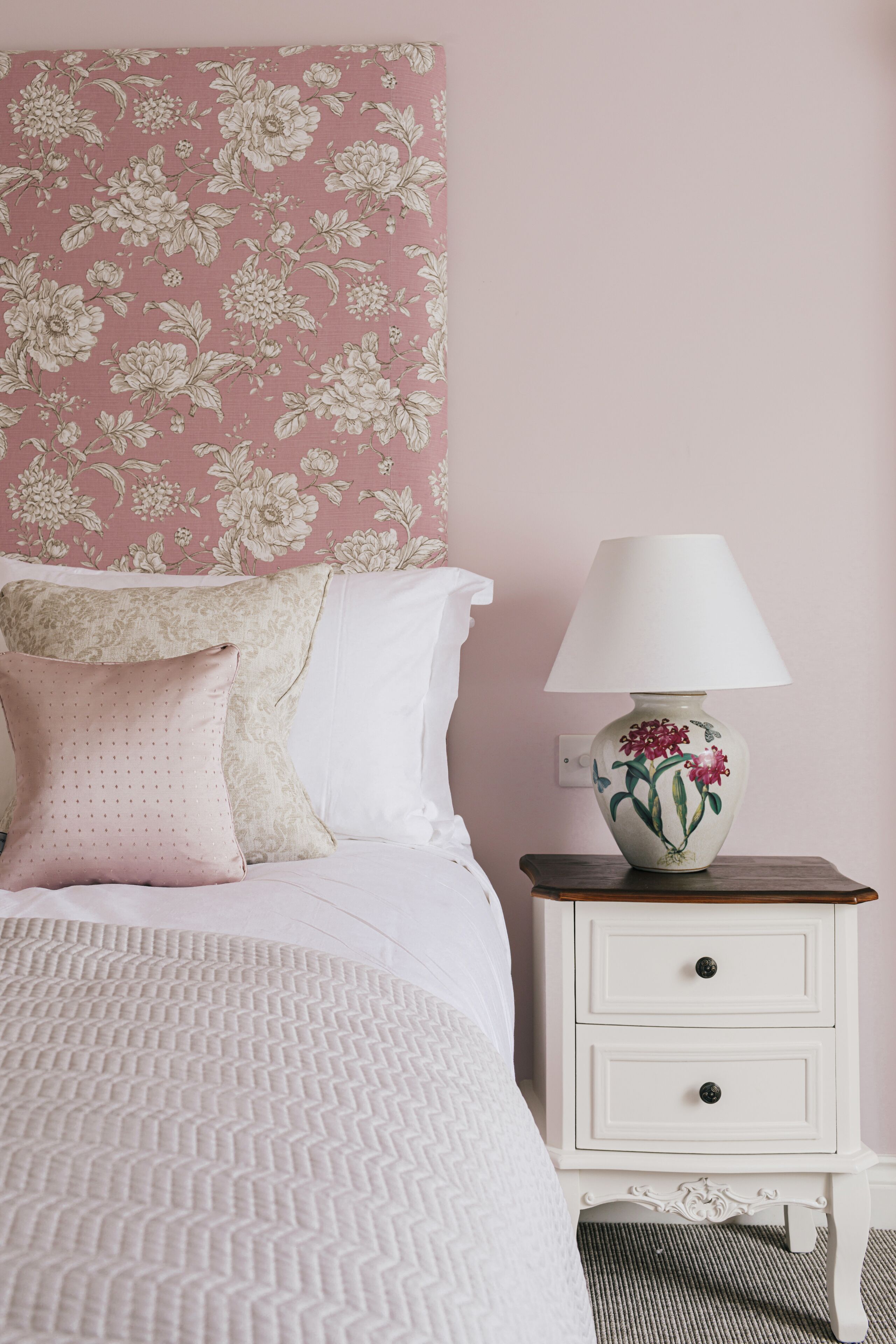 A neatly made bed with a patterned bedspread, a white nightstand with a lamp, and a floral wallpaper on the wall.