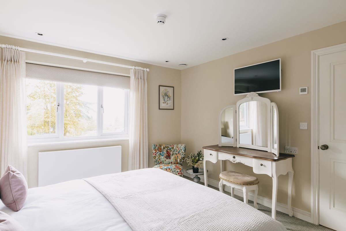Une chambre à coucher comprenant un lit, une commode, une chaise, un téléviseur et une fenêtre avec des rideaux.