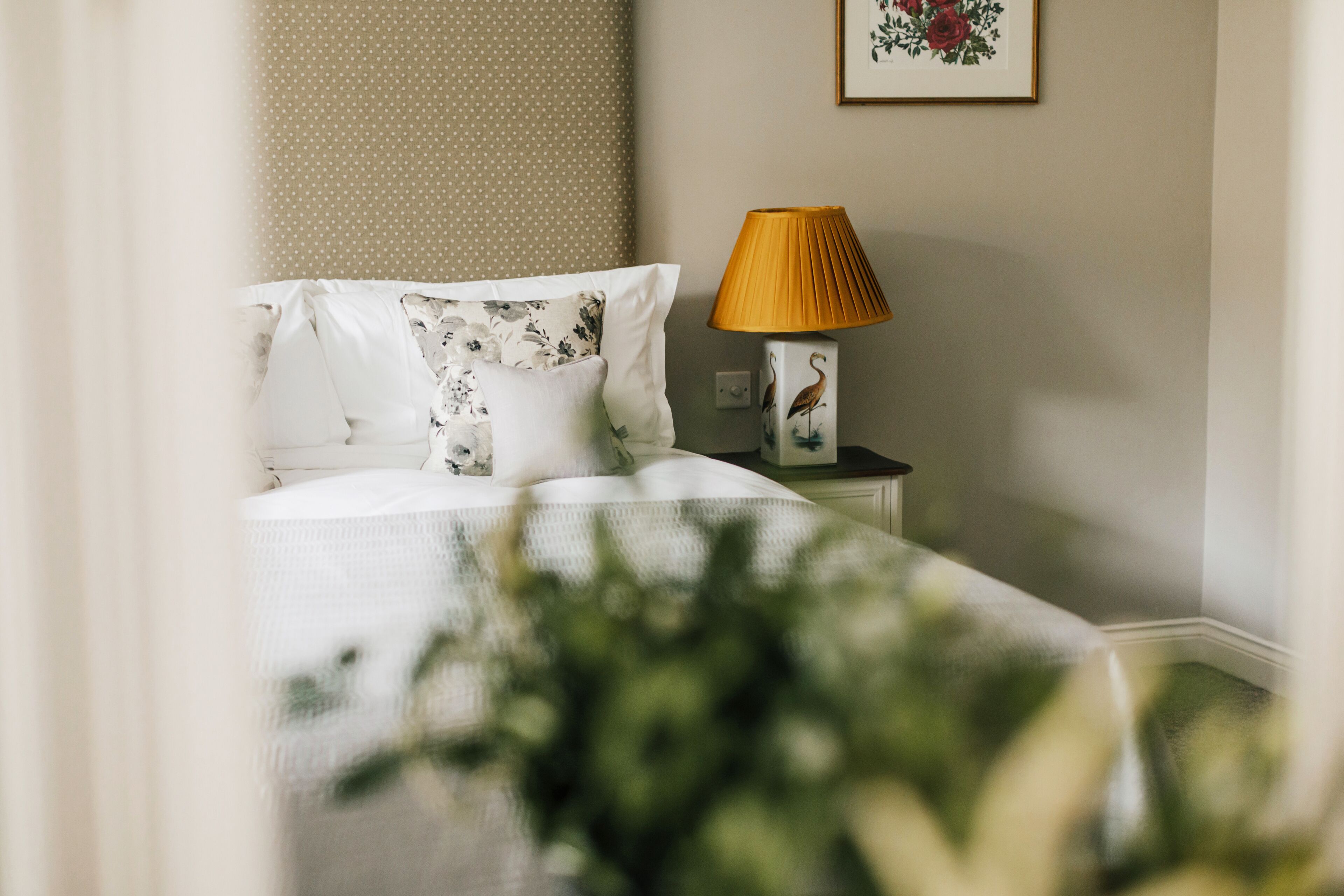 A neatly made bed with white bedding and floral patterns, a bedside table with a lamp featuring a bird design, and a framed floral artwork on the wall.
