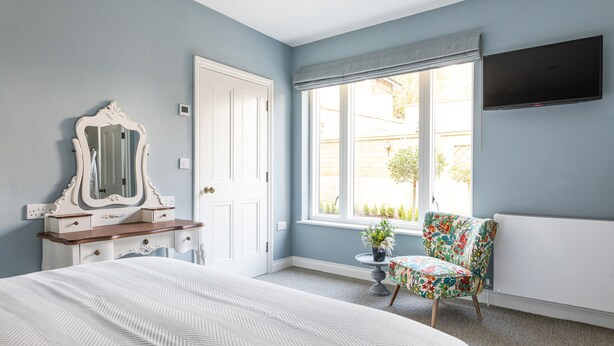 Een slaapkamer met een bed, een dressoir, een spiegel, een raam met gordijnen, een stoel en een televisie aan de muur.