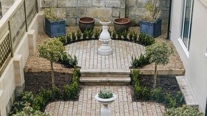 Une cour avec un chemin en briques, des plantes en pot et un pilier central blanc.