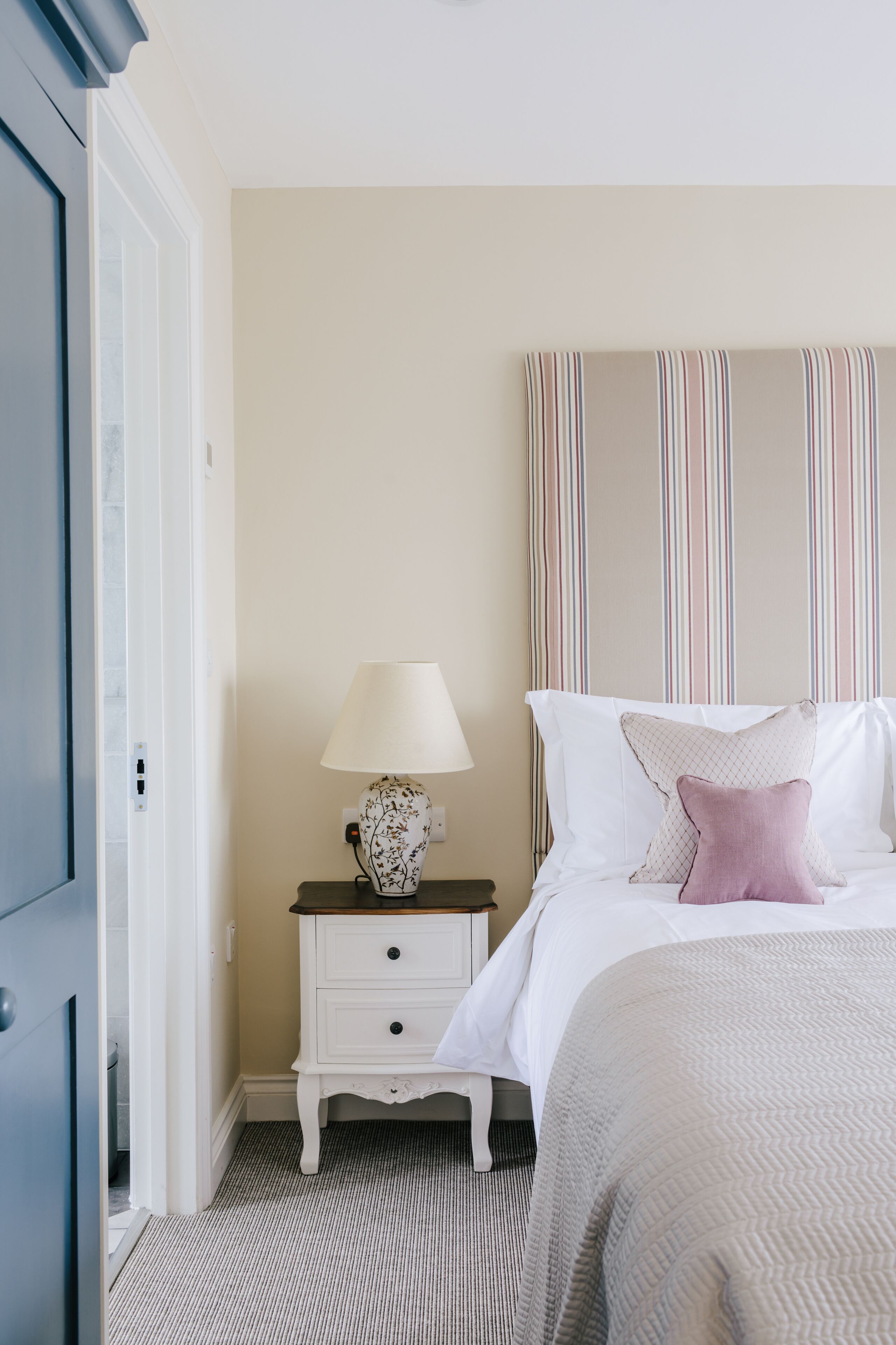 A bedroom with a bed, a nightstand with a lamp, a striped headboard, and a door.
