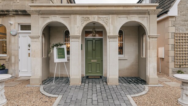 Une entrée d’immeuble avec une porte verte, trois arcades et un petit espace jardin agrémenté d’un panneau indicateur.