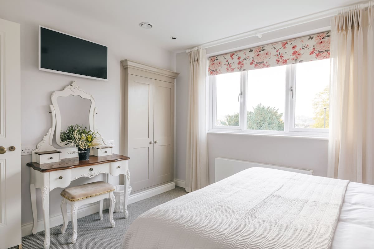 Une chambre à coucher avec un lit, une commode avec un miroir, un banc, une fenêtre avec des rideaux et un téléviseur fixé au mur.