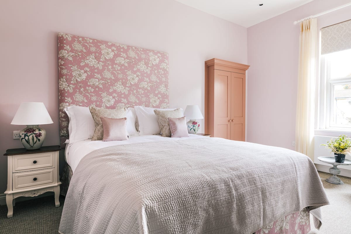 Une chambre à coucher avec une tête de lit à motifs floraux, un lit avec de nombreux oreillers, une table de chevet avec une lampe et une fenêtre avec des rideaux.