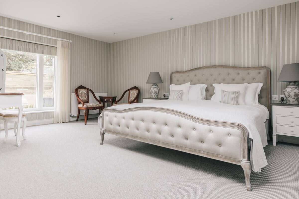 Une chambre à coucher avec un grand lit, deux tables de chevet avec des lampes, une chaise et une fenêtre avec des rideaux.
