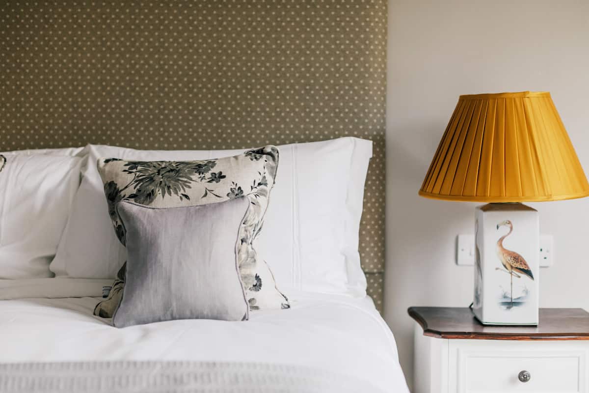 Un lit bien fait, avec du linge de lit blanc et un coussin à motifs floraux, une table de chevet avec une lampe décorée d’un motif d’oiseau, et un mur à pois.