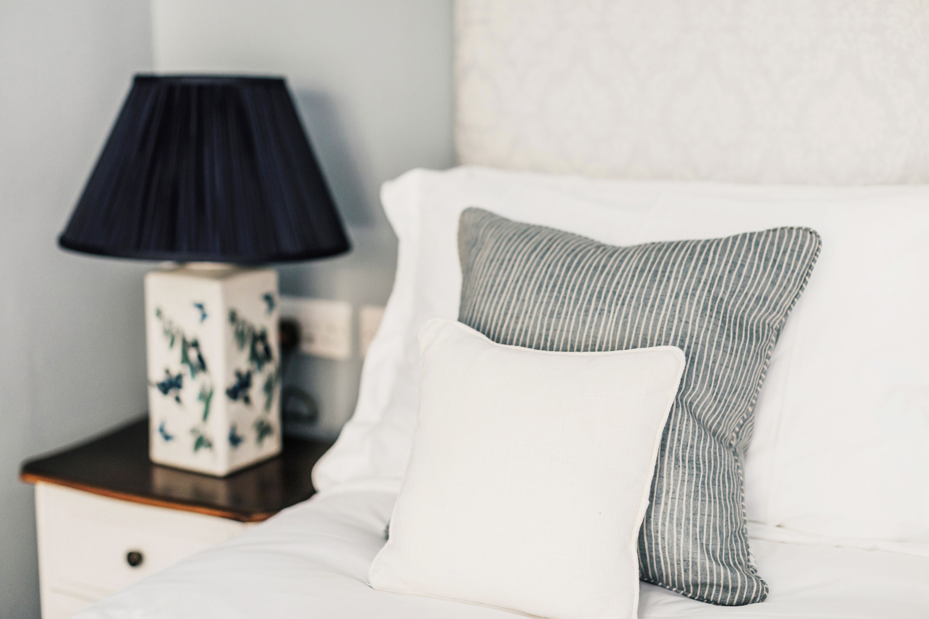 A neatly made bed with white bedding and striped pillows, a dark blue lamp on a wooden nightstand, and a decorative vase with a floral pattern.