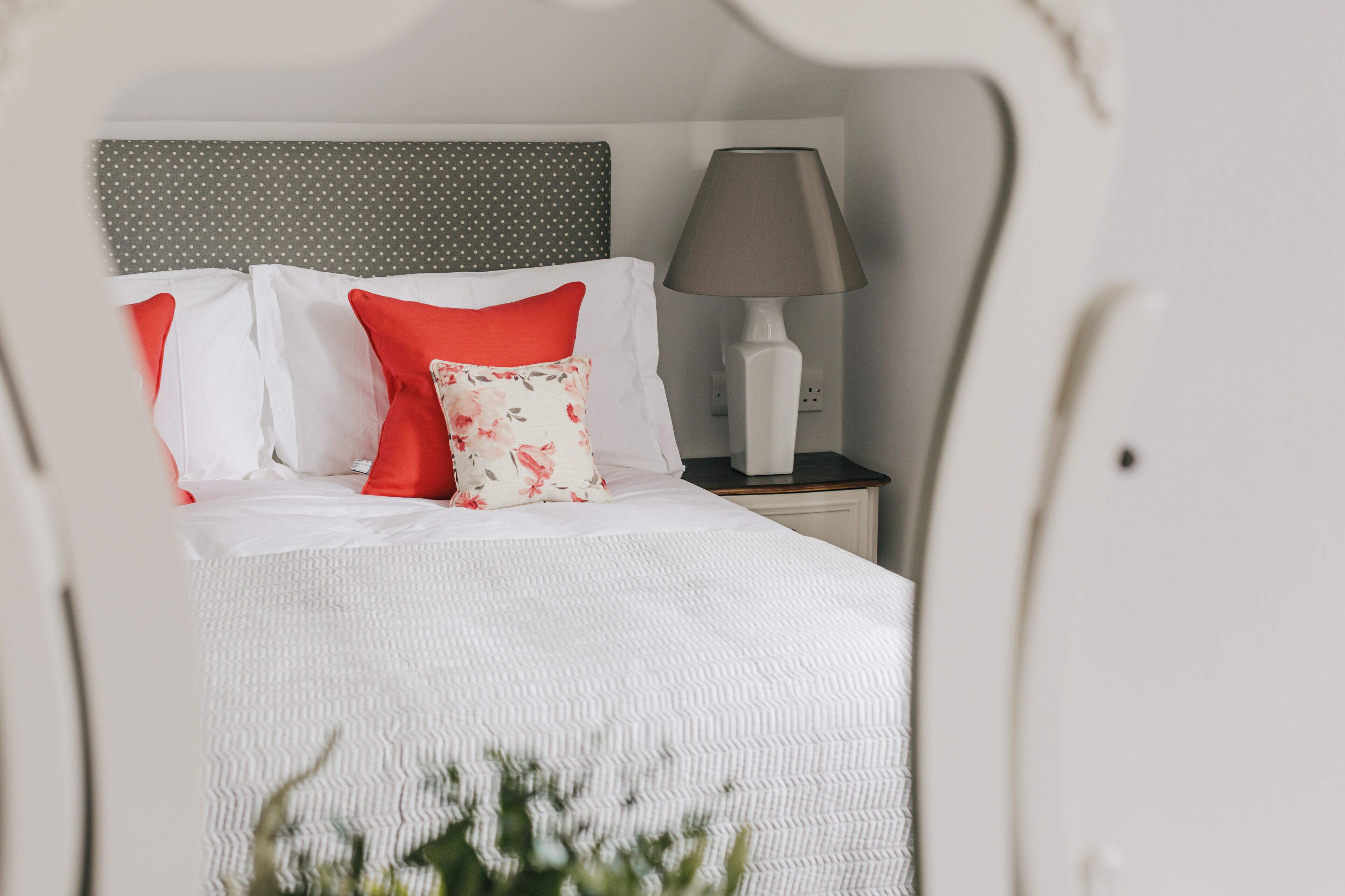 A neatly made bed with white bedding and red pillows, a grey polka dot headboard, and a bedside lamp on a nightstand.
