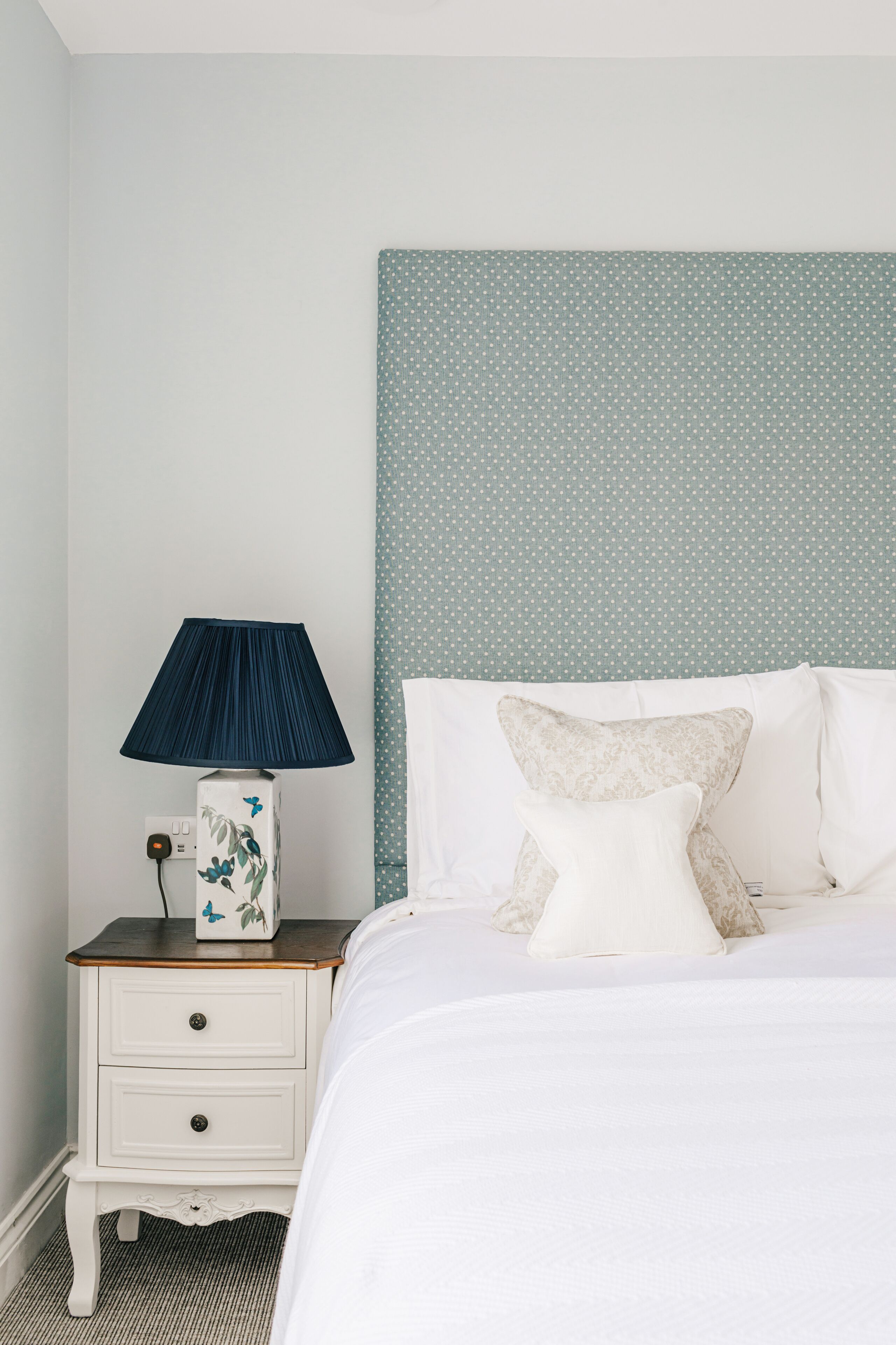A neatly made bed with white linens and multiple pillows, a bedside table with a lamp, and a wall with a polka dot pattern.