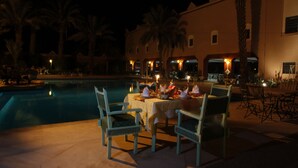 Un área de comedor al aire libre con piscina, palmeras y un edificio al fondo.