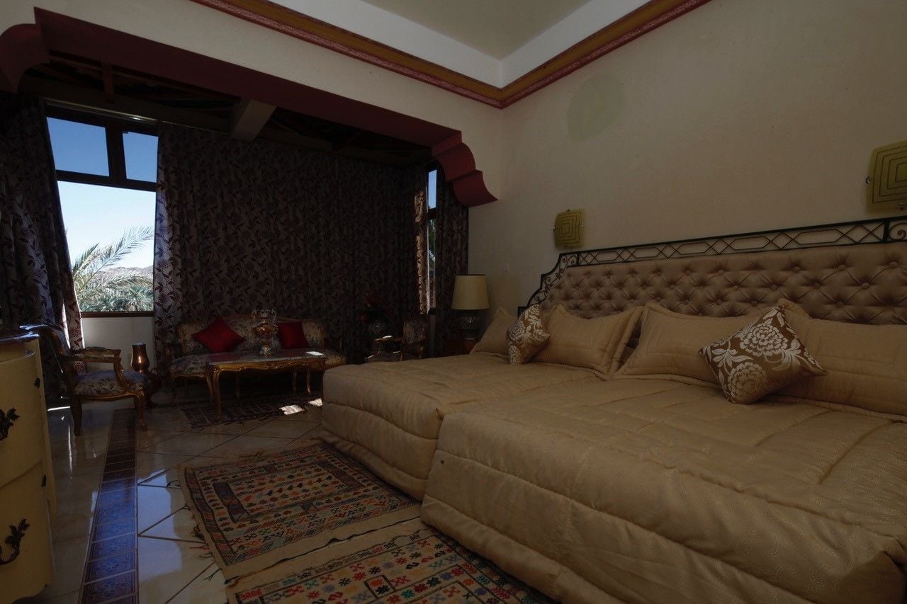 A bedroom with a large bed, a sitting area with a table and chairs, and a view of palm trees through the window.