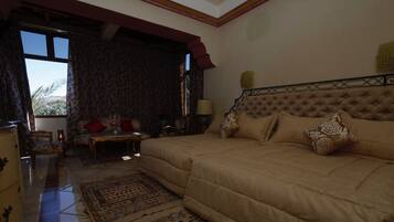 A bedroom with a large bed, a sitting area with a table and chairs, and a view of palm trees through the window.