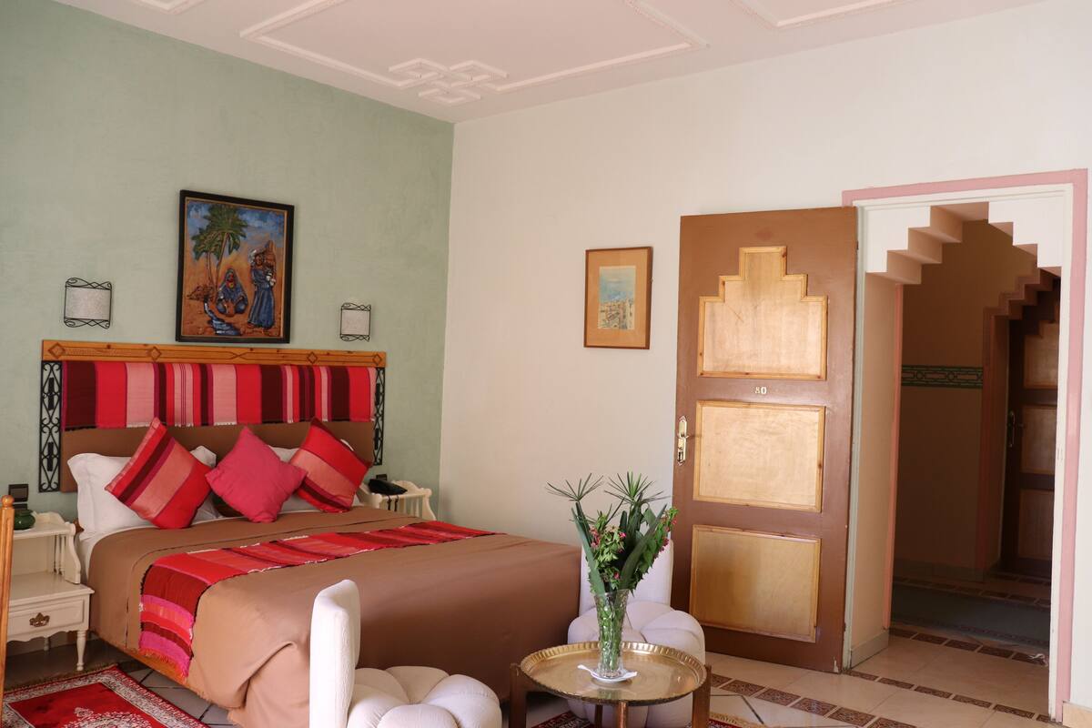 Une chambre d’hôtel avec un lit, une table de chevet, une petite table sur laquelle se trouve un vase, et une porte donnant sur un escalier.