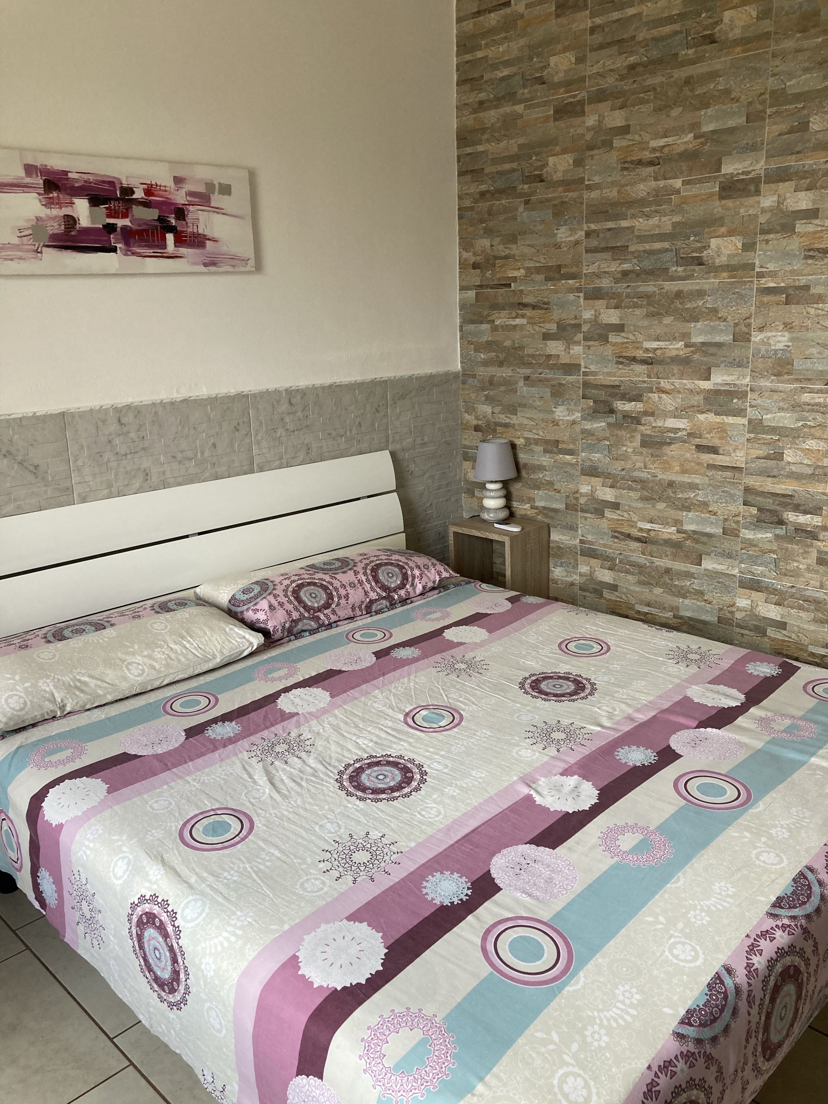 A bedroom with a bed, a nightstand, a lamp, and a stone-patterned wall.