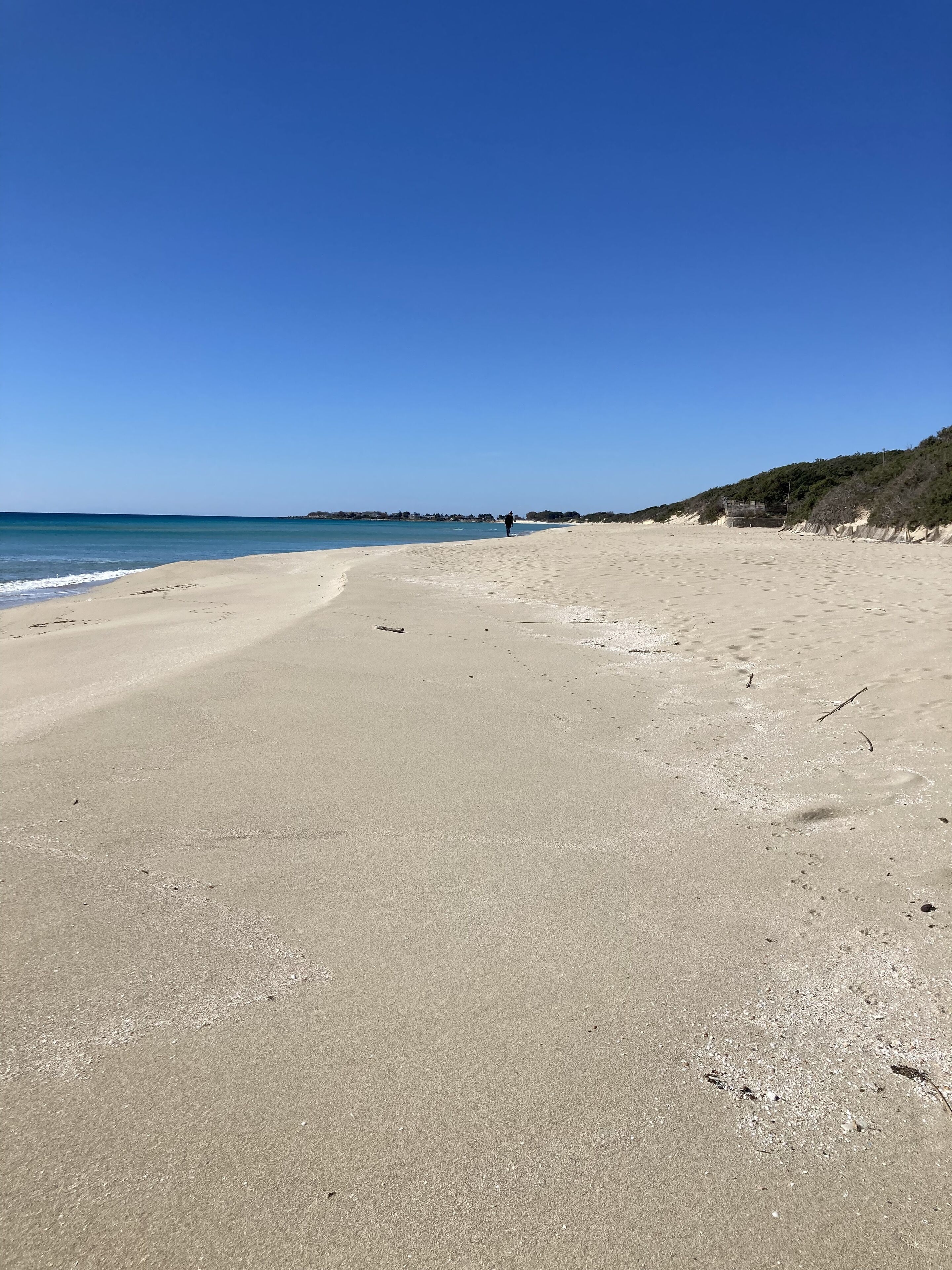 A wide sandy beach with gentle waves, a clear blue sky, and a distant person walking.