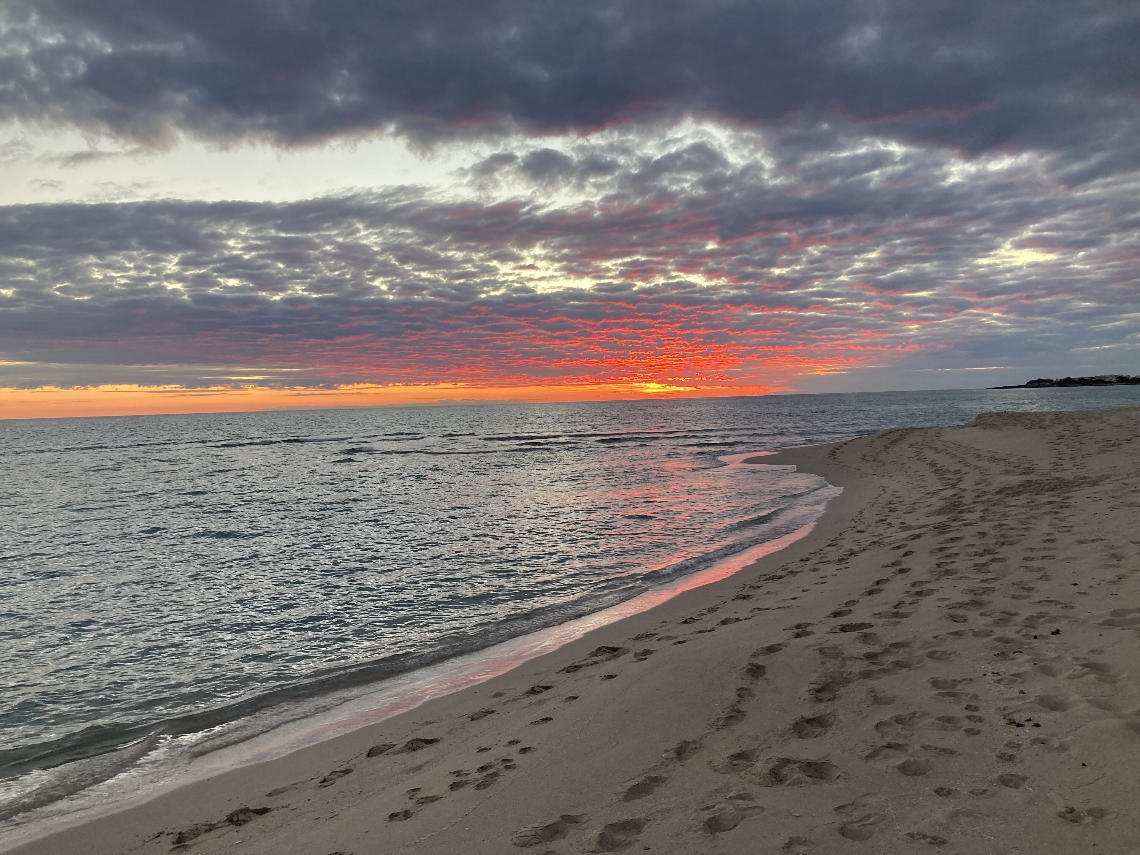 Una playa con huellas que conducen al agua, cielo despejado y un atardecer.