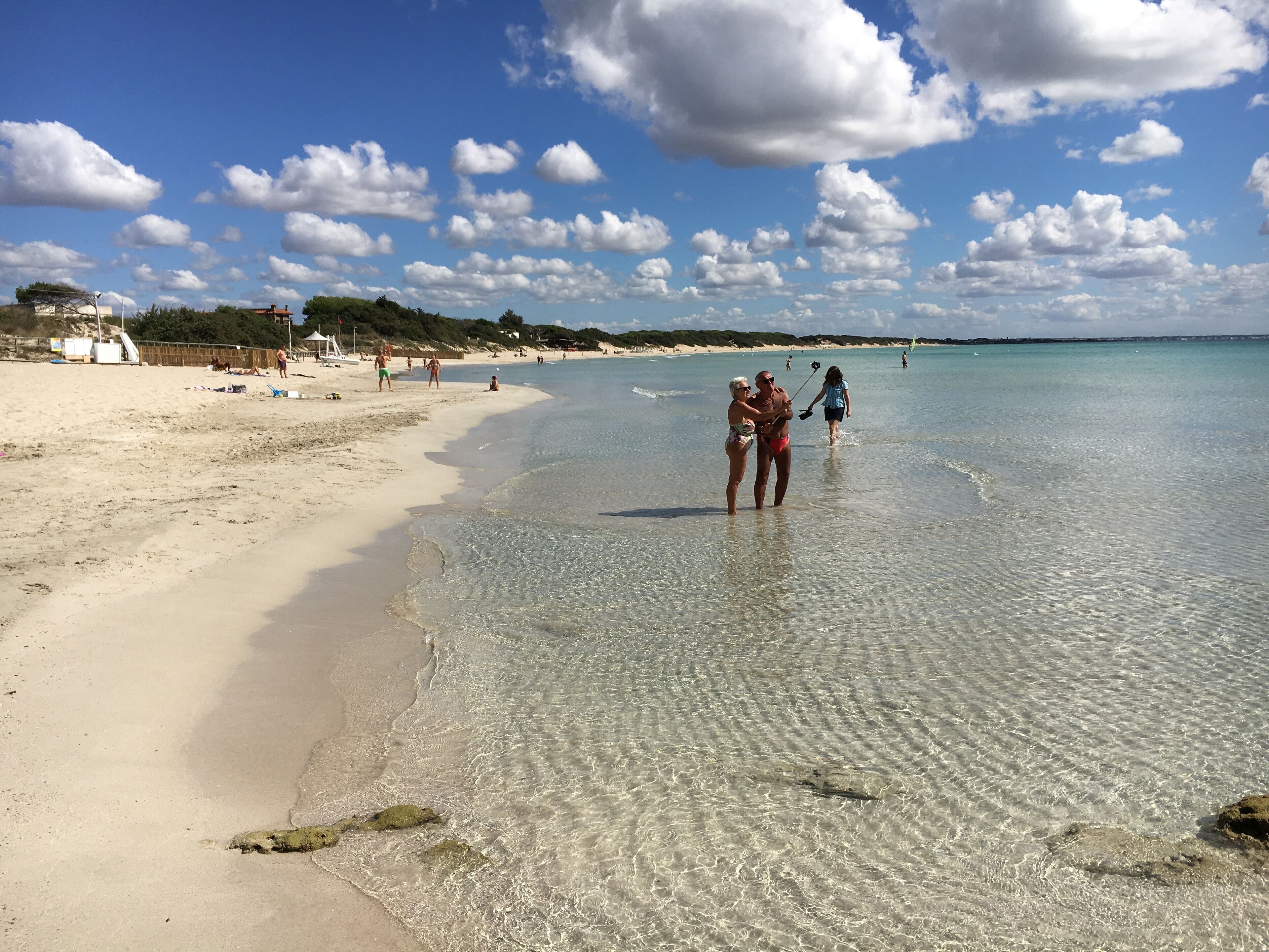 A beach with clear shallow waters, a sandy shore, and a few people walking.